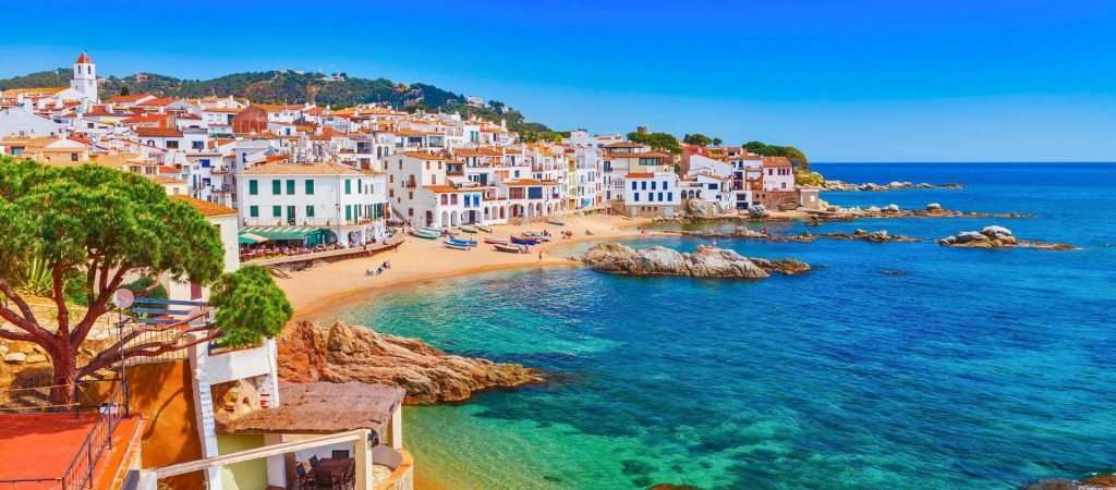 The Best of Spain-This is a picturesque view of a coastal Mediterranean village. It showcases vibrant whitewashed buildings with terracotta roofs, nestled along a sandy beach and rocky shoreline.