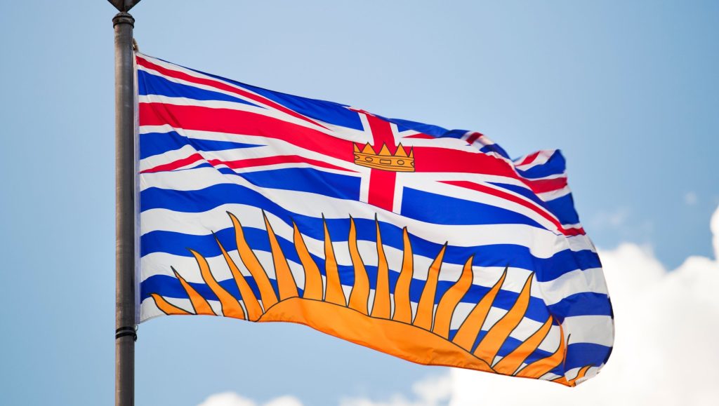 Why 5 Days in BC Made Me Rethink City Life-This image shows the provincial flag of British Columbia, Canada, fluttering in the wind against a bright blue sky with soft clouds. Here's a breakdown of its design and symbolism